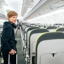 Chłopiec z walizką i słuchawkami na szyi stoi w przejściu samolotu. Zastanawia się, od ilu lat można latać samemu.